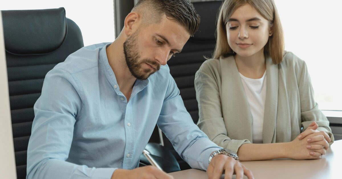 327 A Caucasian man in a blue button-up shirt writing something down with a pen, while a Caucasian woman wearing a beige blazer looks at what he's writing.