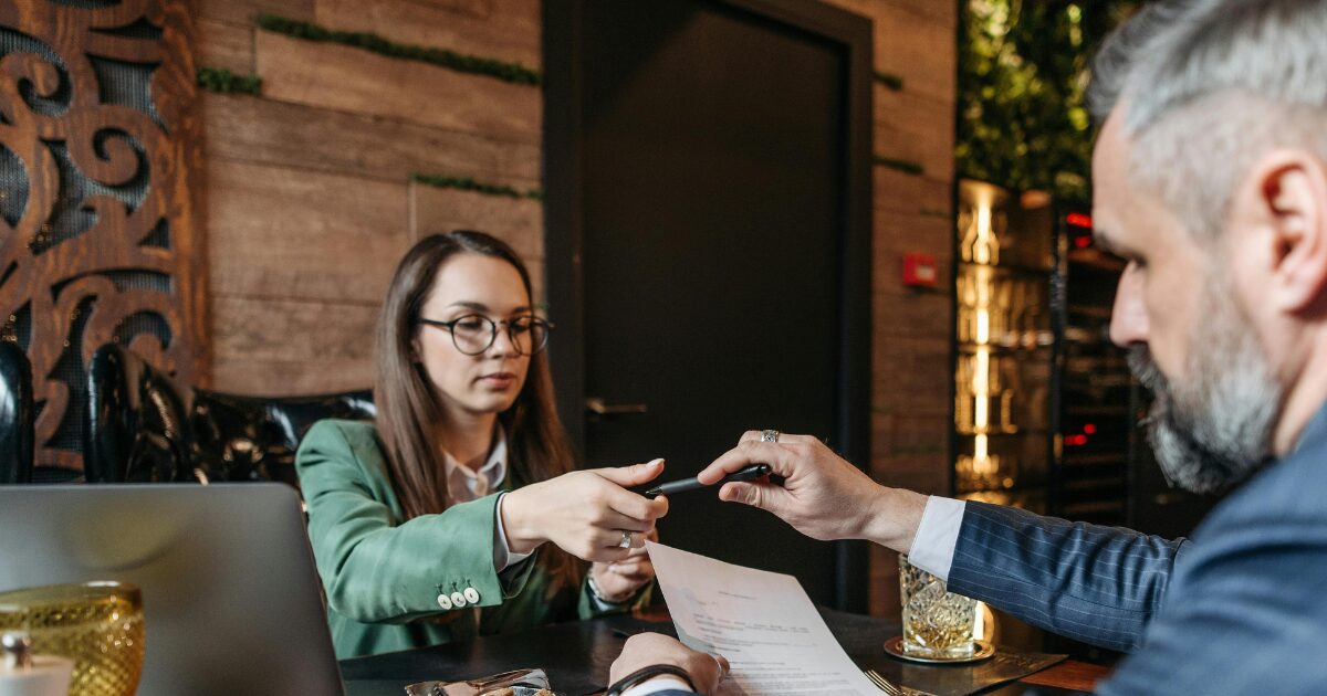 328 Caucasian woman sitting across from a middle-aged Caucasian man, as he passes her a pen while he holds a piece of paper.