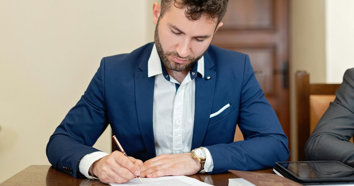 330 A Caucasian man in a blue blazer, sitting at a wood table, writing something on a piece of paper.
