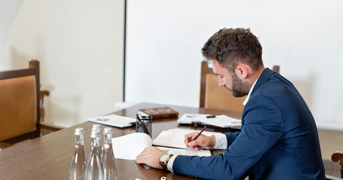 331 A Caucasian man in a blue blazer, sitting at a wood table, holding a pen while writing something on a document.