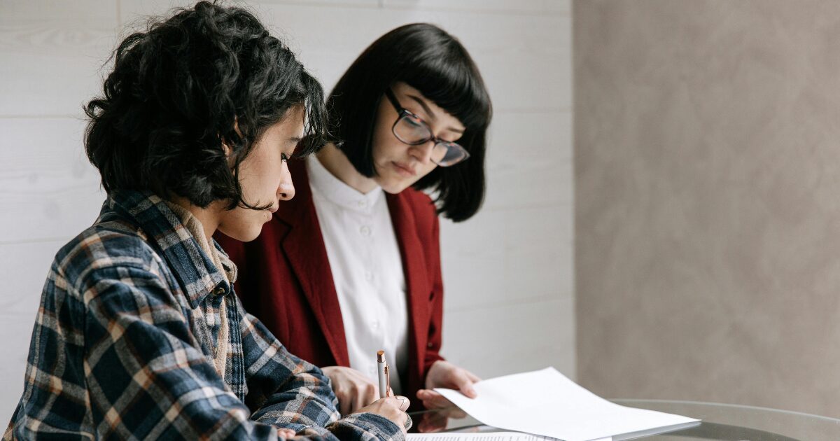 332 A Latin woman wearing a plaid shirt is signing a document, while a Caucasian woman wearing a red blazer watches her sign