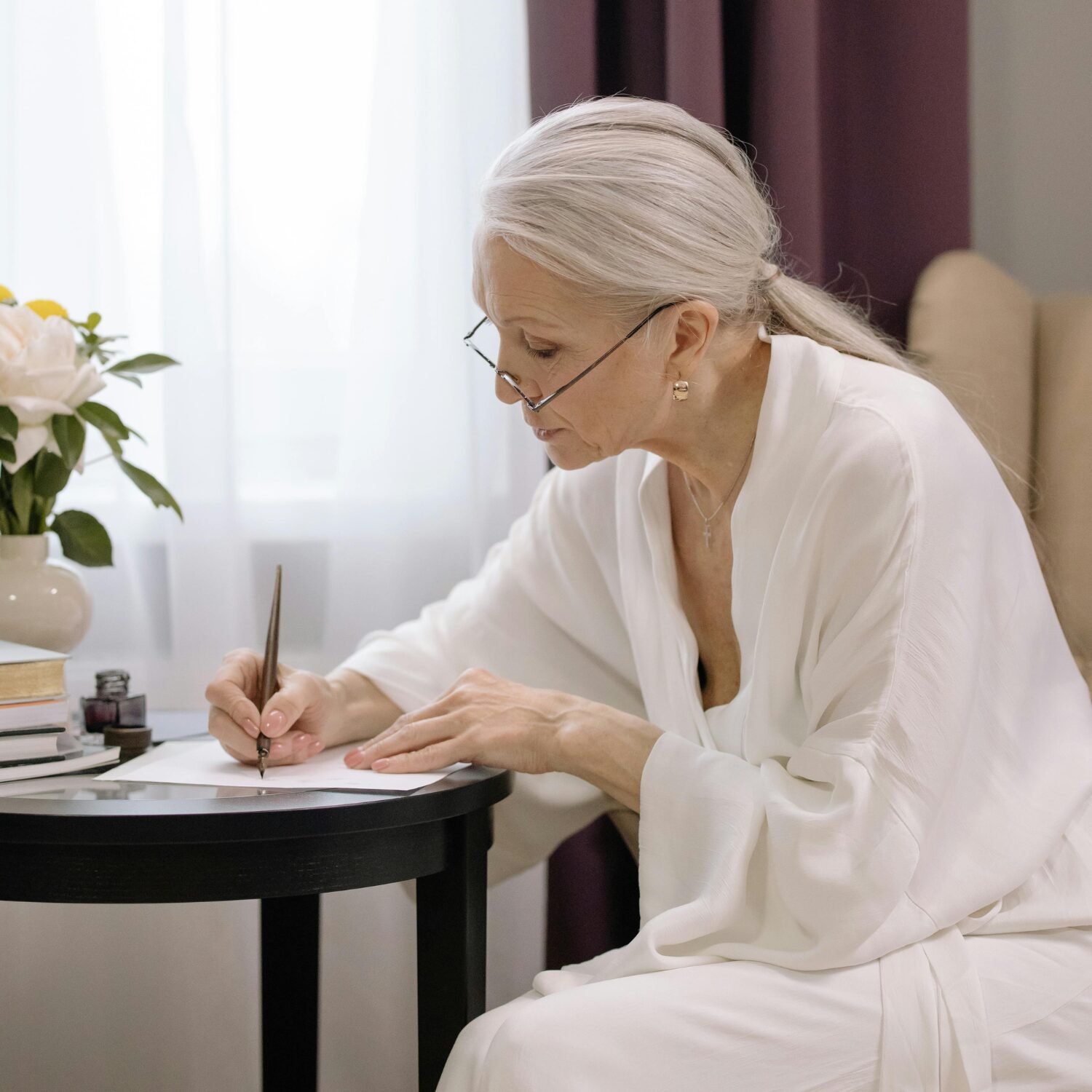 Senior Caucasian woman in a white dress, sitting at a dark brown table, signing a piece of paper.