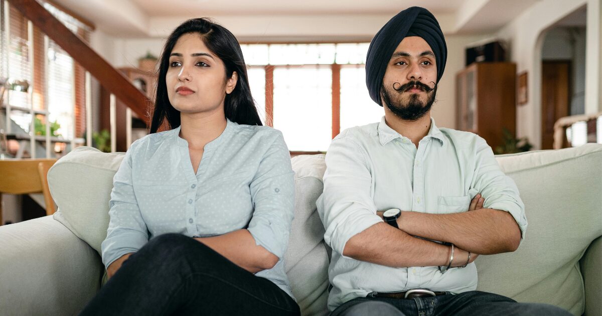 193 An East Indian male and female couple, sitting side by side on a couch, looking sad with their arms crossed. Both are wearing button-up shirts.
