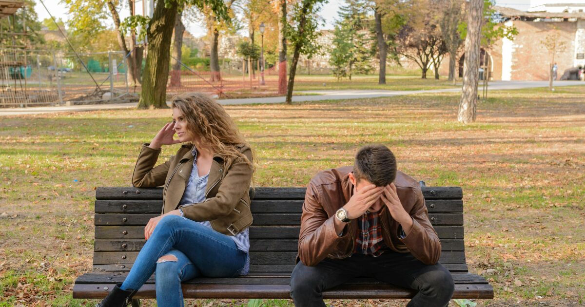 194 A Caucasian male and female couple sitting on a park bench in the fall. He has his head in his hands and she is looking off to the side, both seem sad.