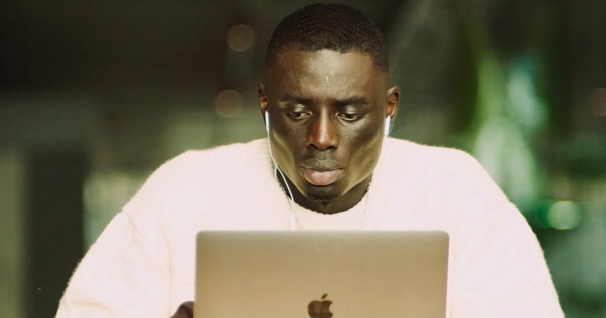 196 Black Man wearing a white shirt and headphones, looking at his open MacBook laptop.