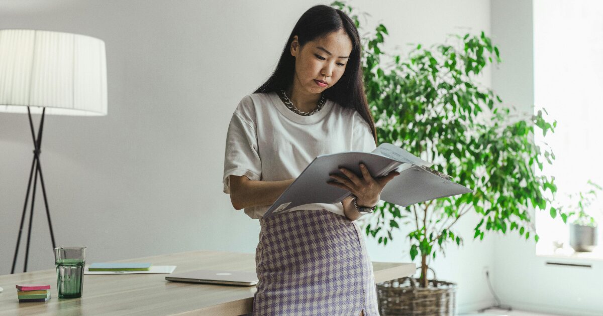 An East Asian woman standing in an office, looking over legal documents, with a plant to her left. 