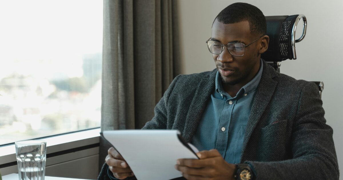 Black man in a blazer, sitting at a desk looking at legal documents.