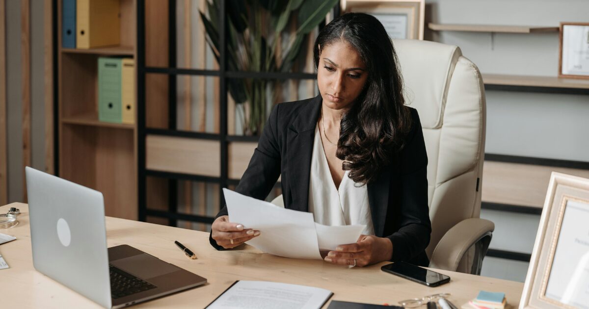 A Black woman wearing a black blazer sitting at a wood desk, holding a document in each hand, while looking over them.