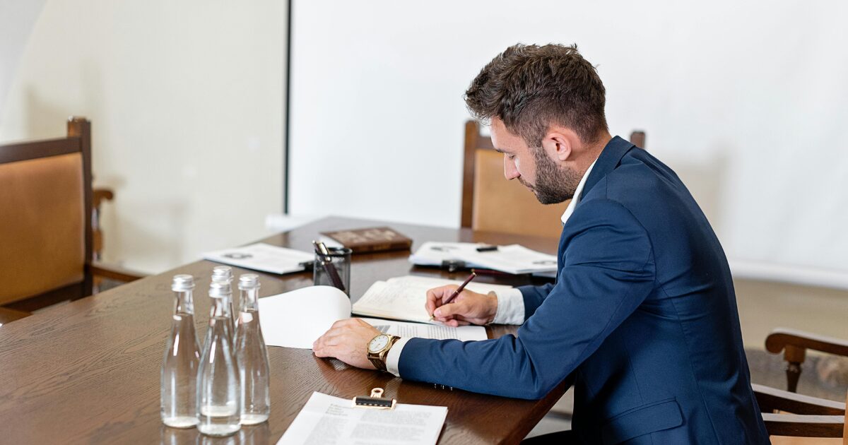 278 A Caucasian man wearing a blue blazer, sitting at a wood desk looking over paperwork with a pen in his hand.