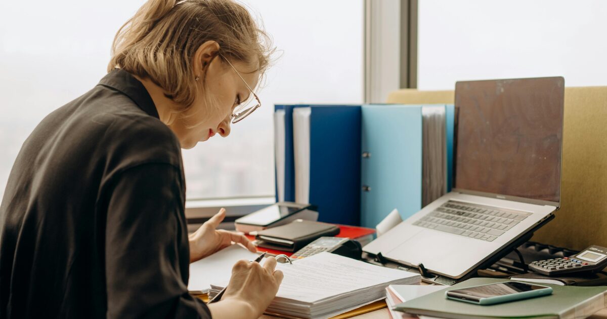 280 a Caucasian woman wearing a dark brown blazer, sitting at a desk while looking over paperwork in front of her.