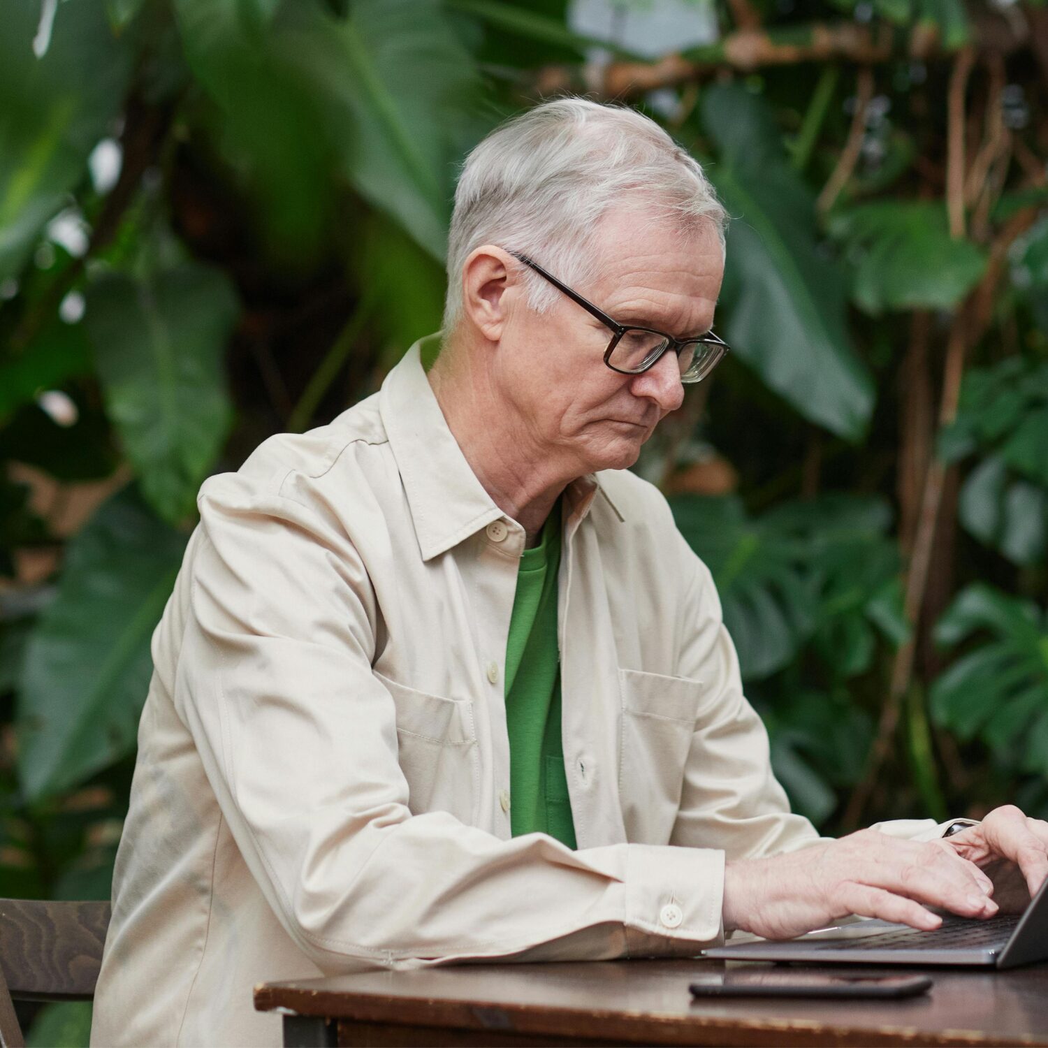 A senior Caucasian man sitting at a desk on his laptop with green plants in the background.