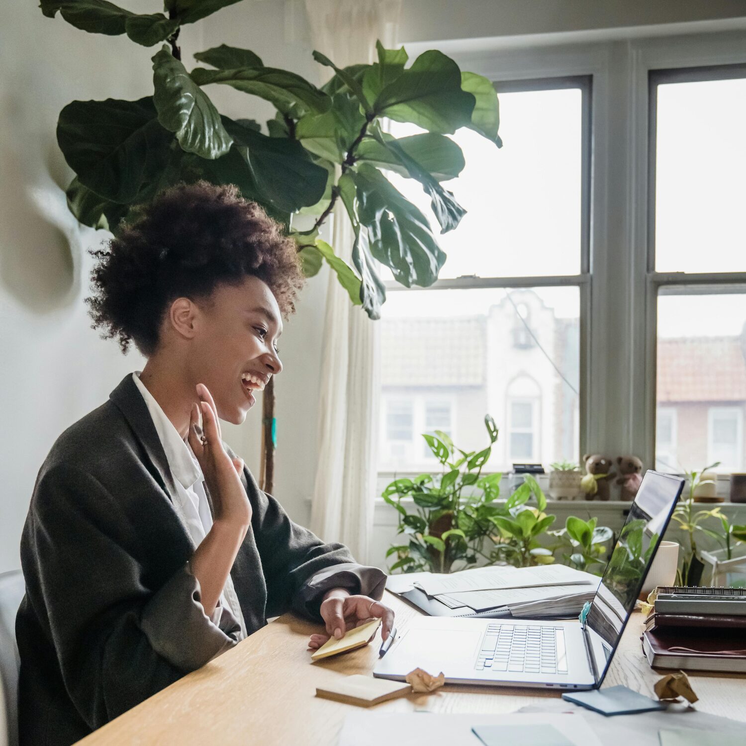 A young Black woman in a dark gray blazer, smiling on a video call on her laptop. She is holding her hand up as if she is declaring an oath.