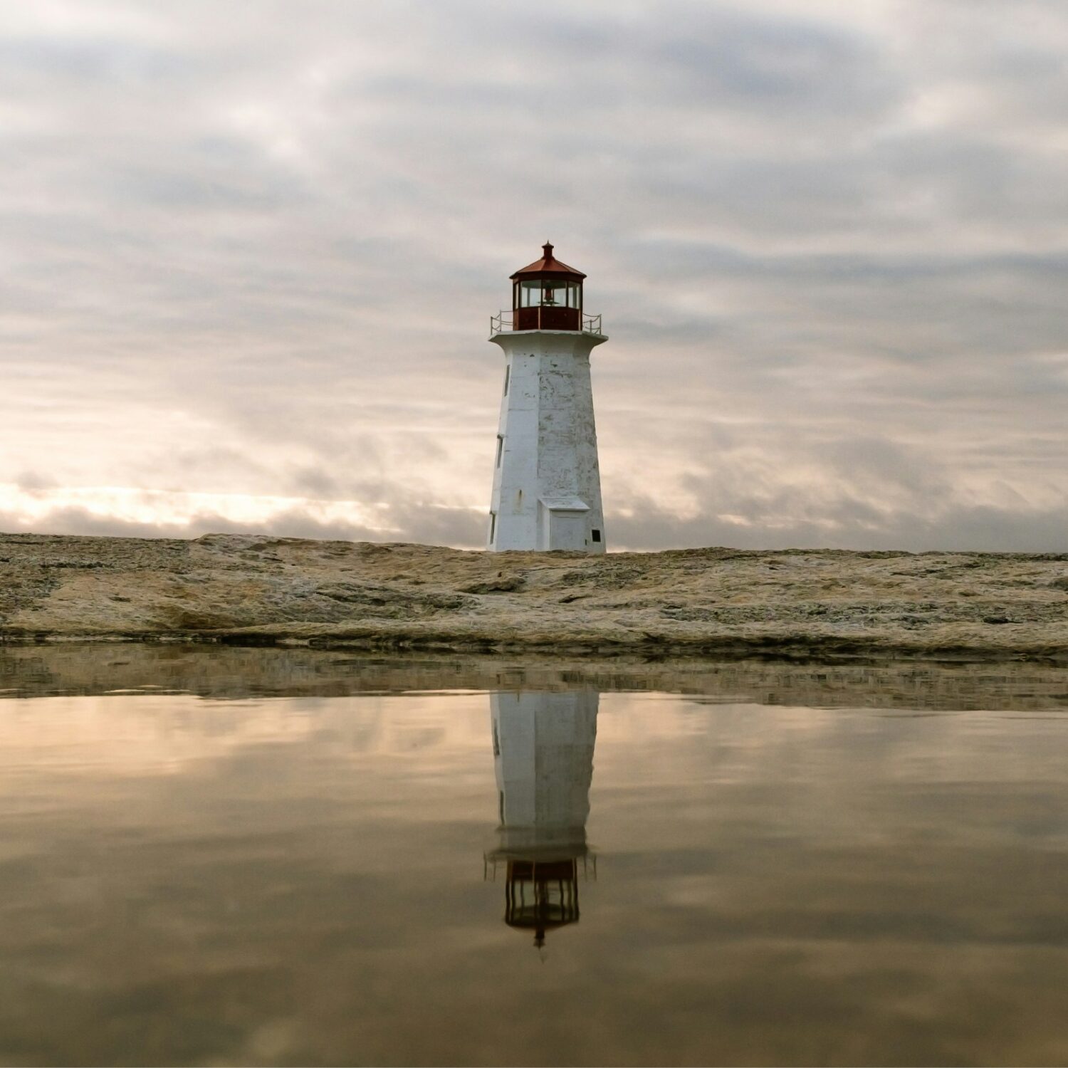 A white lighthouse on land with grey water in front of it and a cloudy sky above.