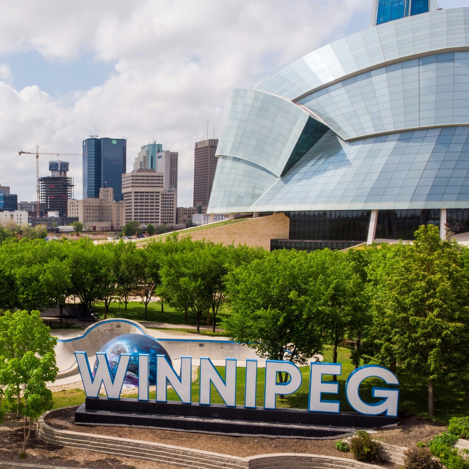 A large white sign that says Winnipeg in big capital letters. There are trees and buildings in the background.