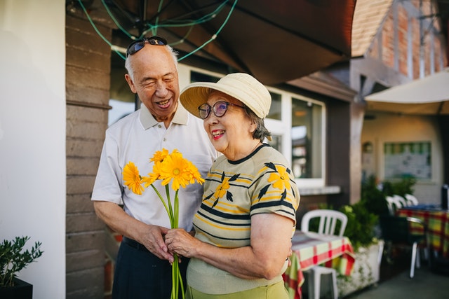 An elderly East Asian couple in front of a cottage. They are wearing summer clothing and she is holding yellow flowers.
