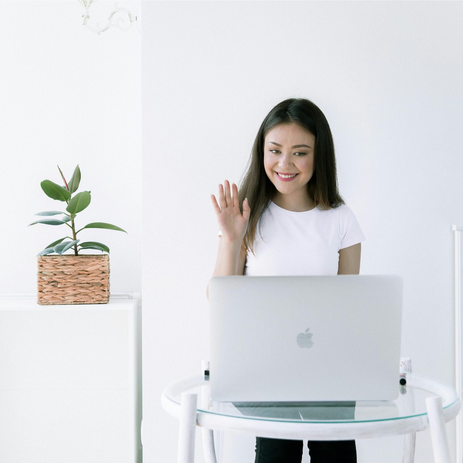 A young East Asian woman wearing a white t-shirt, waving to the camera on her laptop during a video call.
