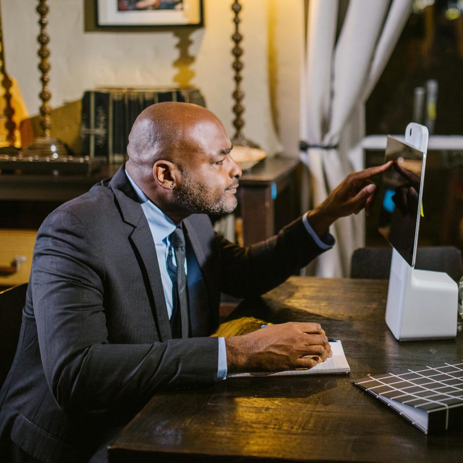A Black, middle-aged man sitting at a desk wearing a black suit. He is using a tablet.