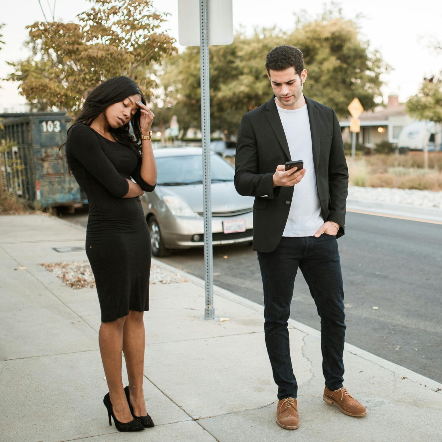 A Black woman standing beside a Caucasian man on the street. He is on his phone, she looks upset while looking down.