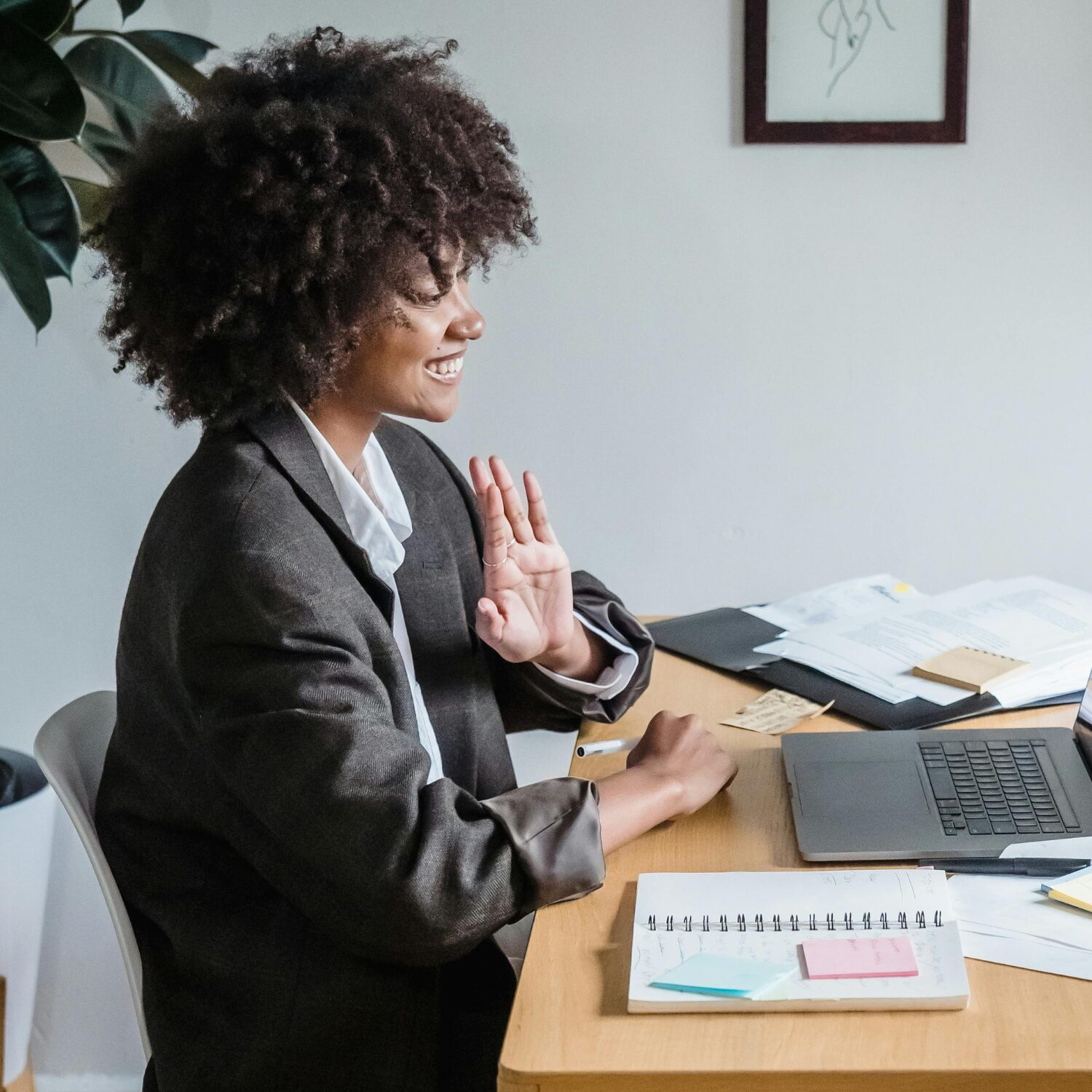 A young Black woman sitting at a wooden desk, wearing a brown blazer. She is on a video call, holding her hand up as if giving an oath.