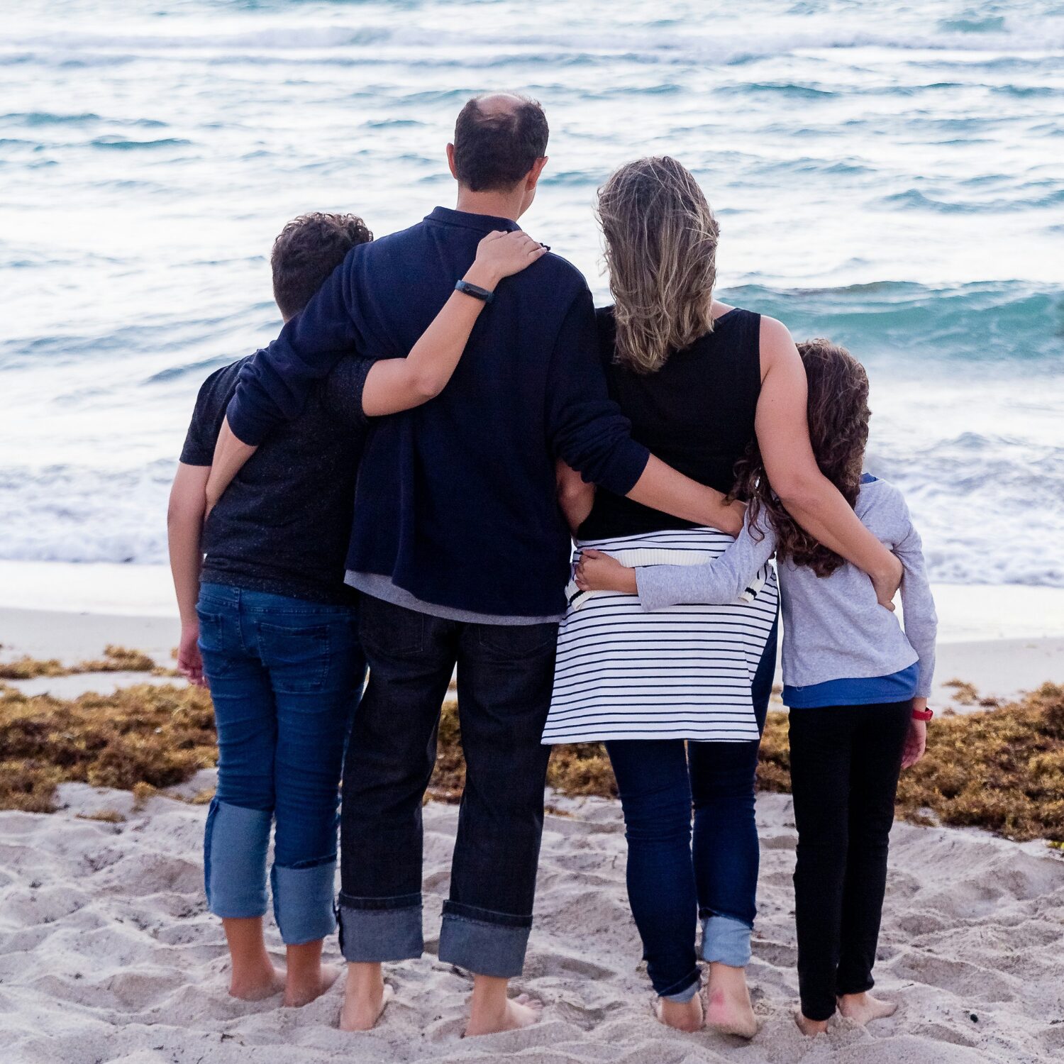 A family of four consisting of a mom, dad, son, and daughter. The kids are young and the family is standing barefoot in sand, facing the ocean with their arms around each other. Their backs are to the camera.