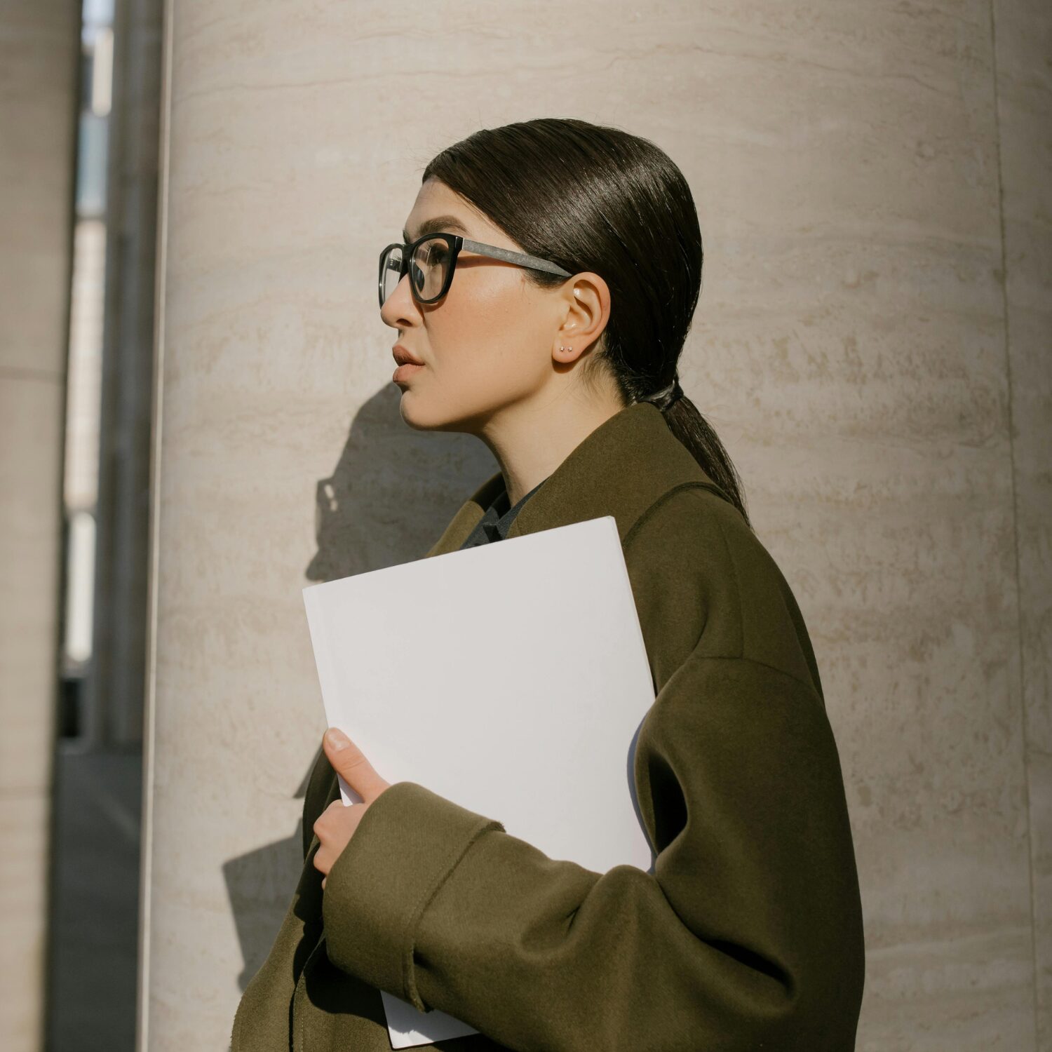 Young Caucasian woman wearing eyeglasses and an olive green coat, holding a document in her hand.