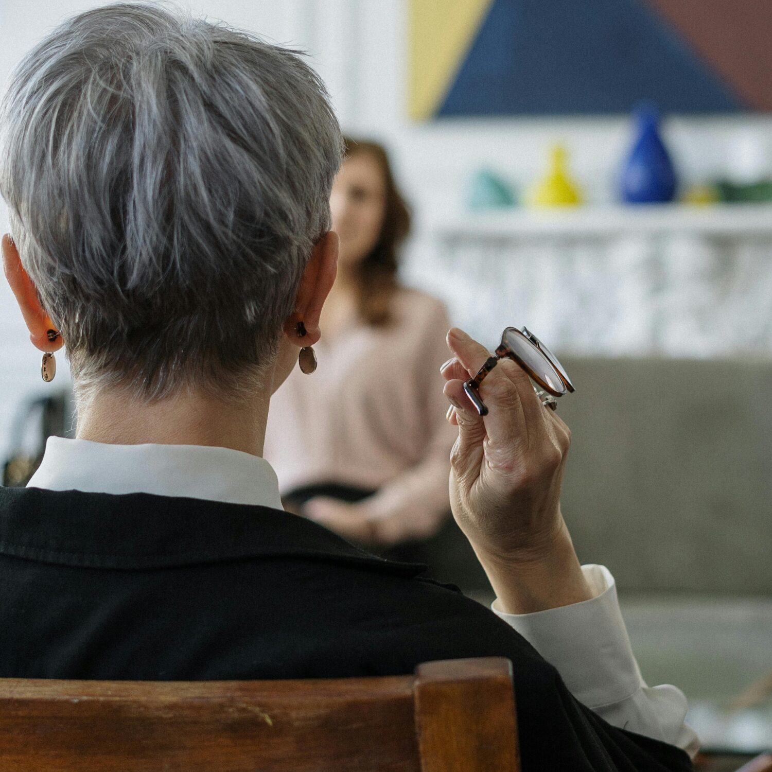 A woman with short hair facing away from the camera, looking at a woman sitting across from her, who is sitting on a grey couch.