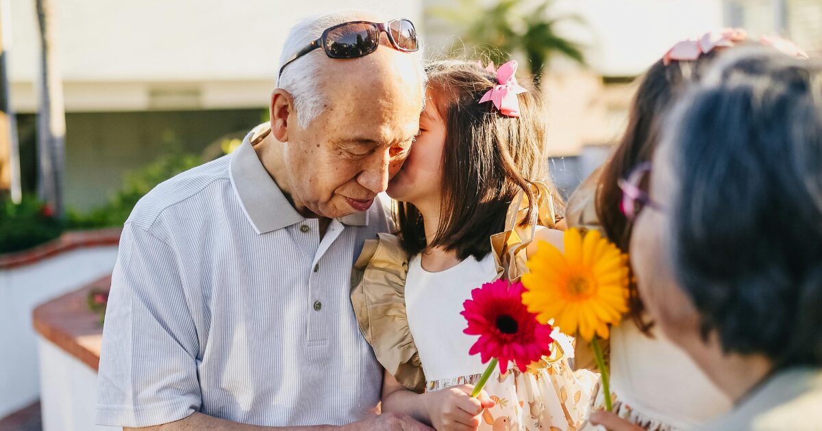 East Asian grandpa holding his young granddaughter, who is kissing his cheek while holding flowers.