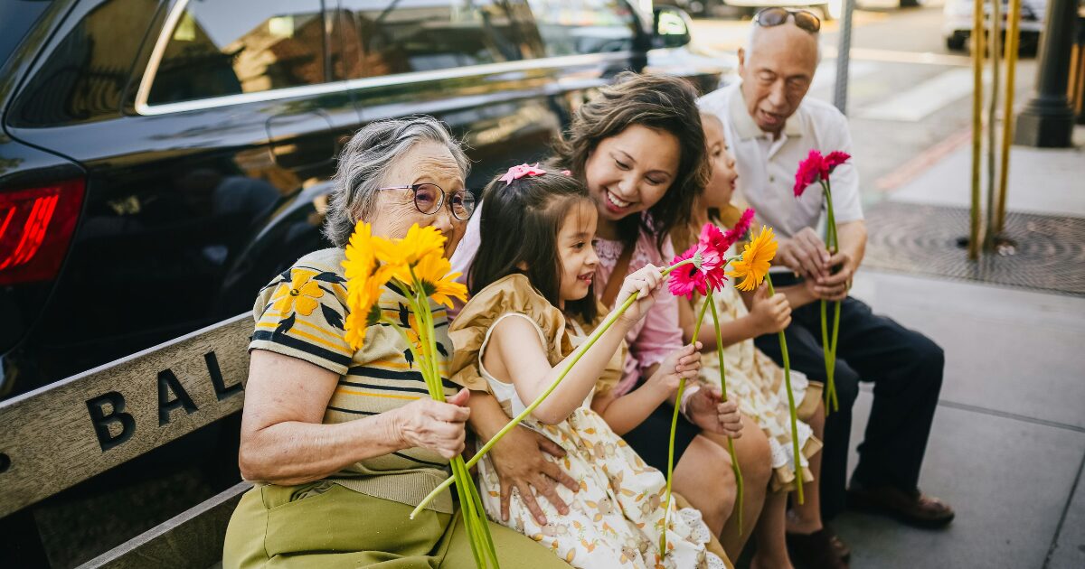 East Asian grandparents invited to visit Canada, spending time with their adult daughter and grandaughter, sitting on a bench holding flowers.