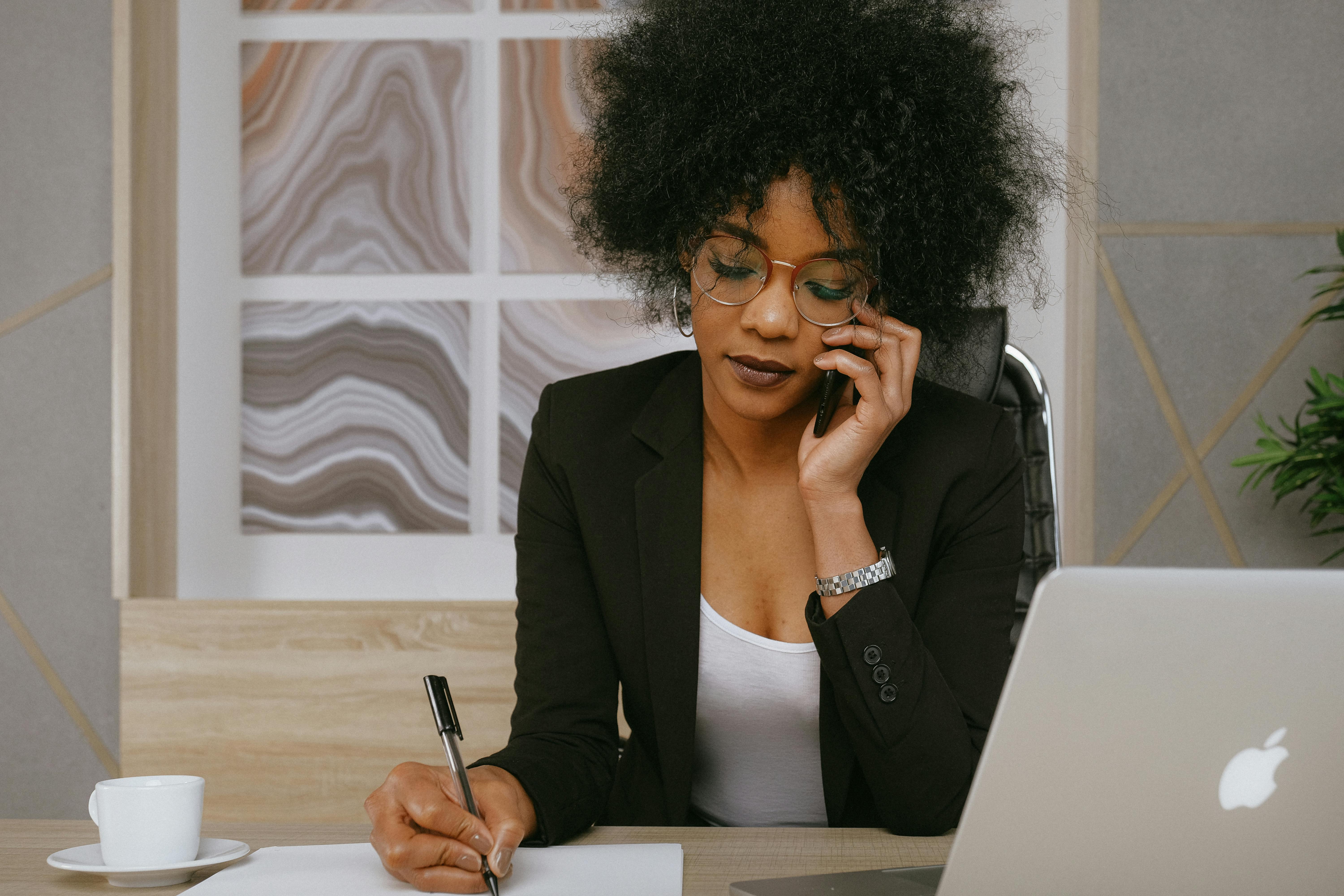 Young Black woman writing something on a piece of paper while on the phone. She is wearing a white shirt with a black blazer.