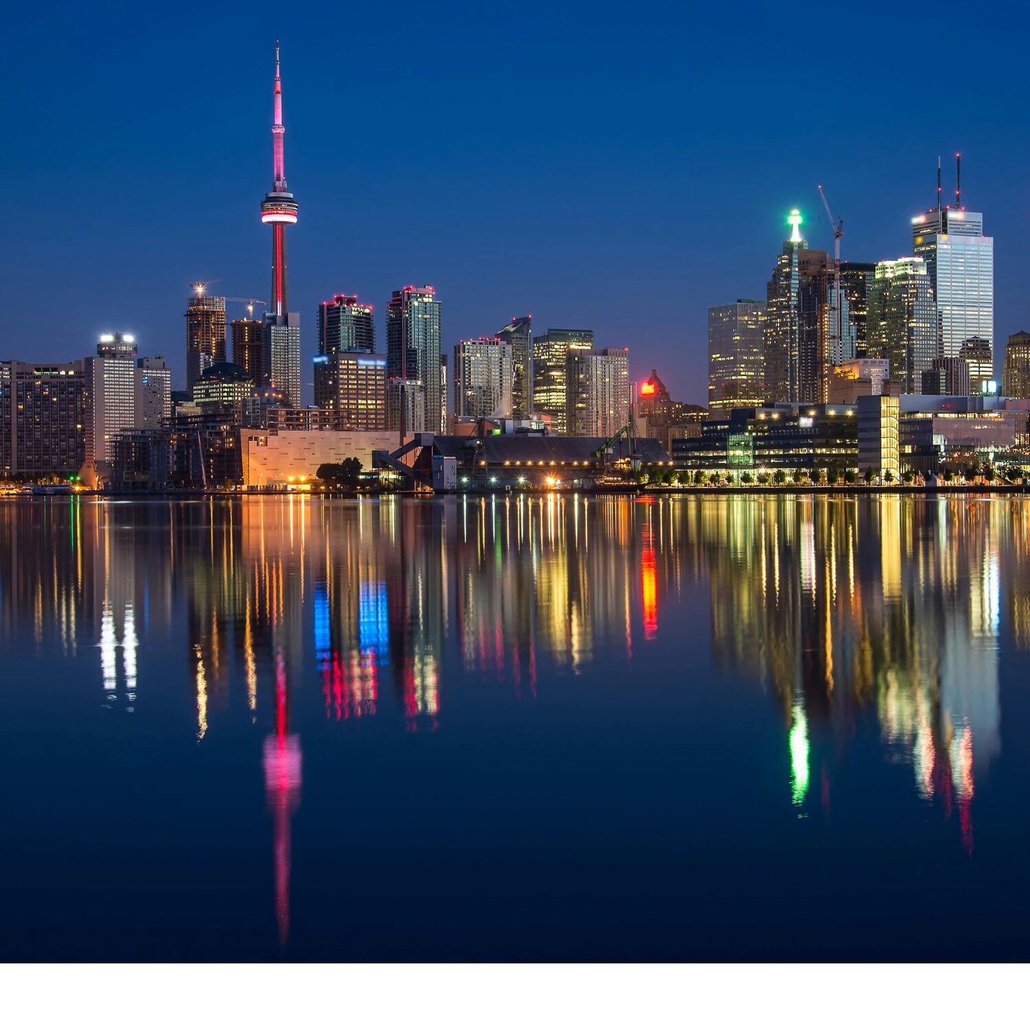 Toronto skyline in the evening in front of a large body of water. You can see the reflection of the skyline in the water.
