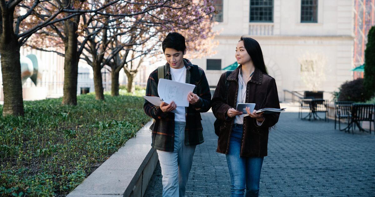 A Caucasian man wearing a checkered coat and an East Asian woman wearing a brown coat walk together outdoors. The man flips through papers while the woman looks at him.