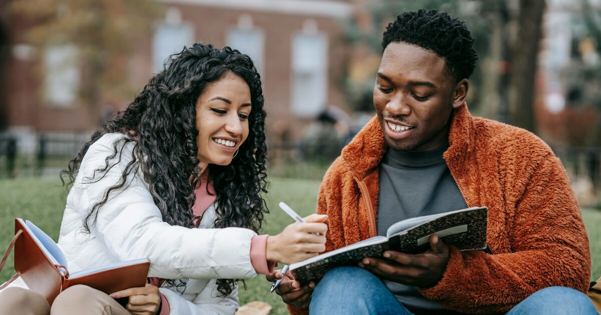 A Latina woman wearing a white jacket and a Black man wearing an orange coat sits together outdoors. The man is holding a notebook while the woman writes something to it. 