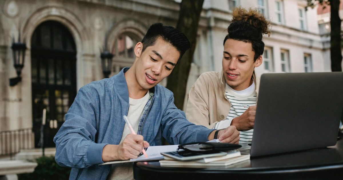 Two Caucasian men sit together outdoors, one in a denim blue jacket writing in a notebook while the other, in a beige coat, looks at it.