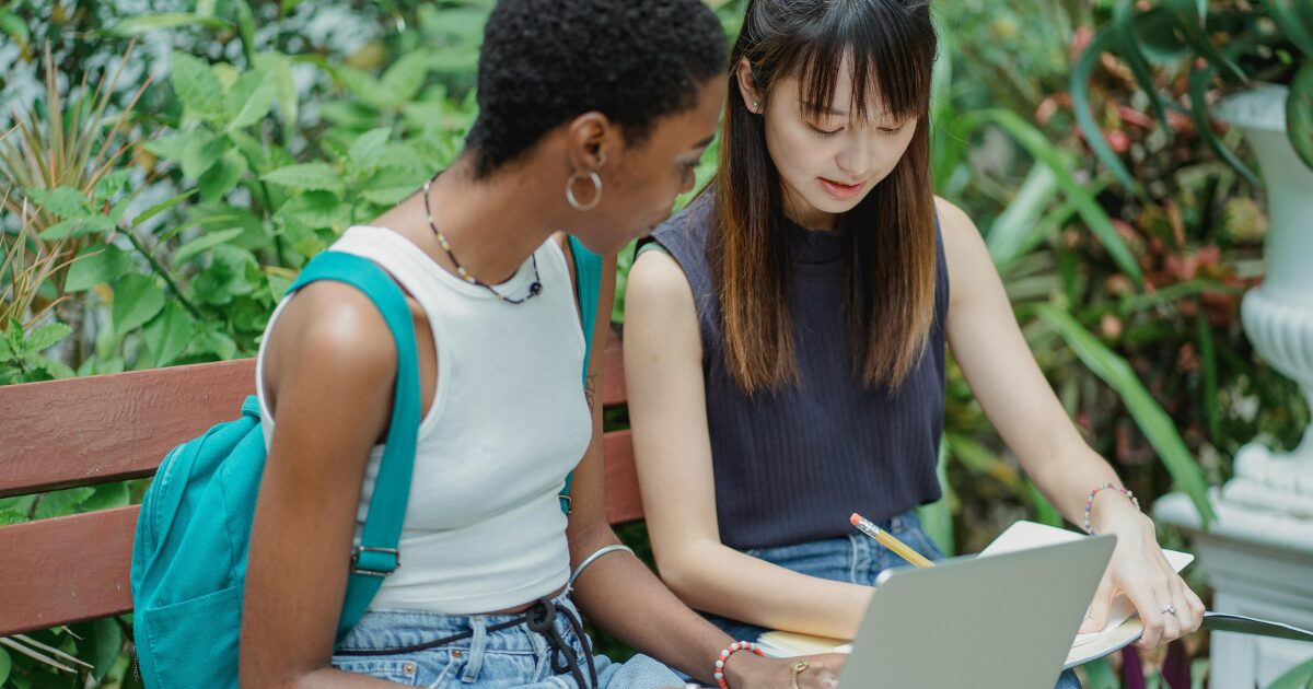 A Black woman in a white sleeveless top sits with an East Asian woman in a dark blue top. The East Asian woman writes in a notebook while the Black woman, holding an open laptop on her lap, looks at her.
