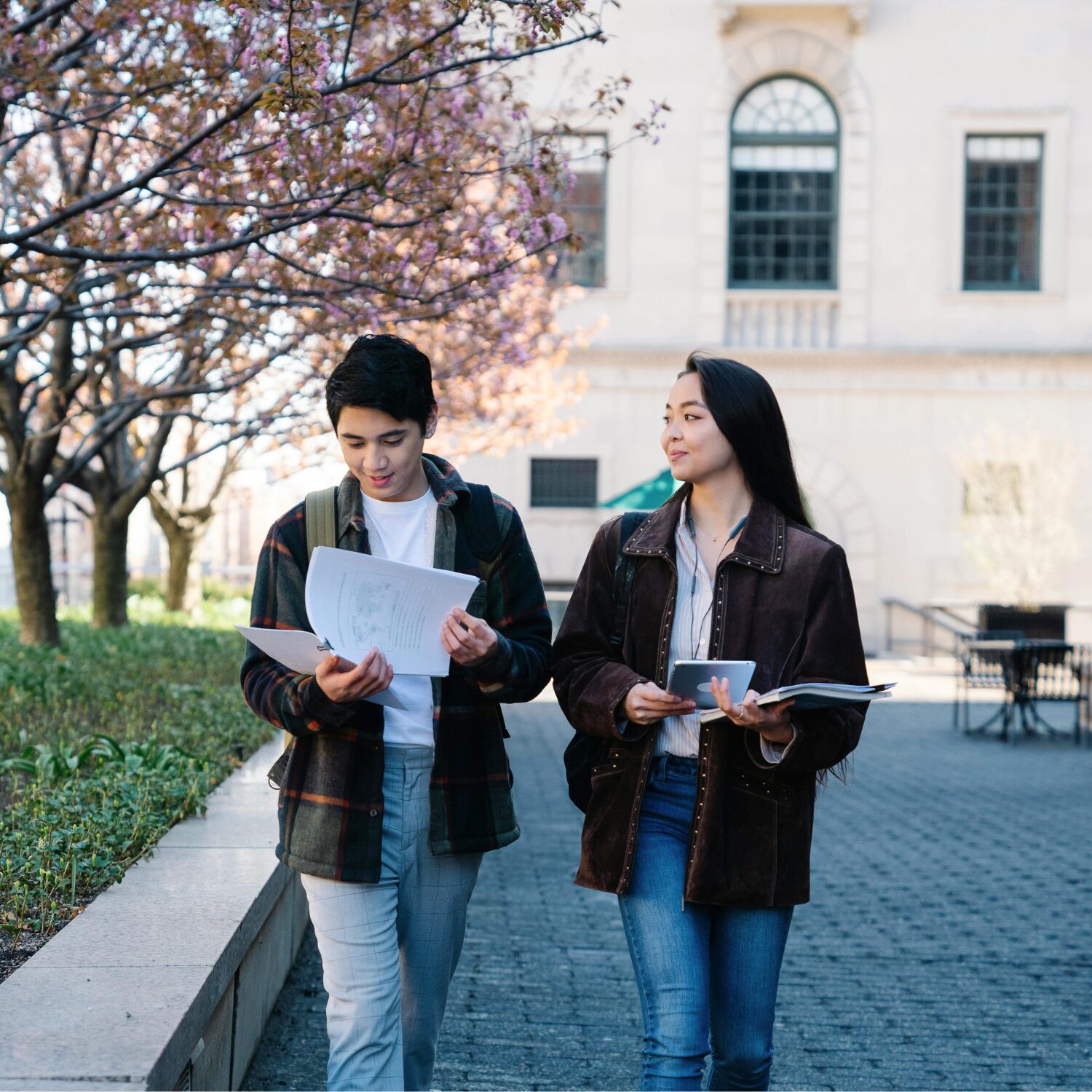A Caucasian man wearing a checkered coat and an East Asian woman wearing a brown coat walk together outdoors. The man flips through papers while the woman looks at him.