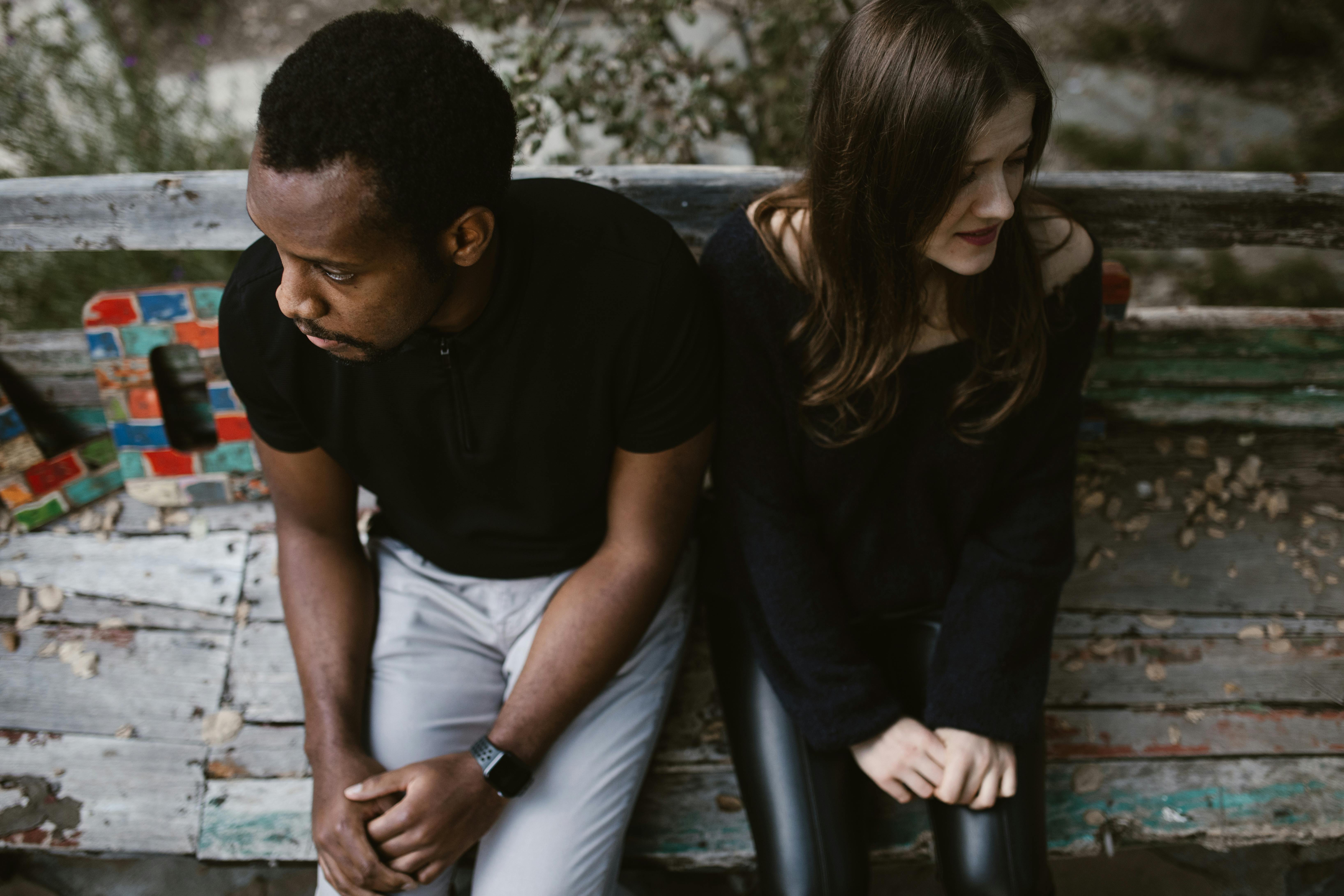 A couple sitting outside on a wood bench, looking in opposite directions, looking upset. The couple is a Black man and a Caucasian woman.
