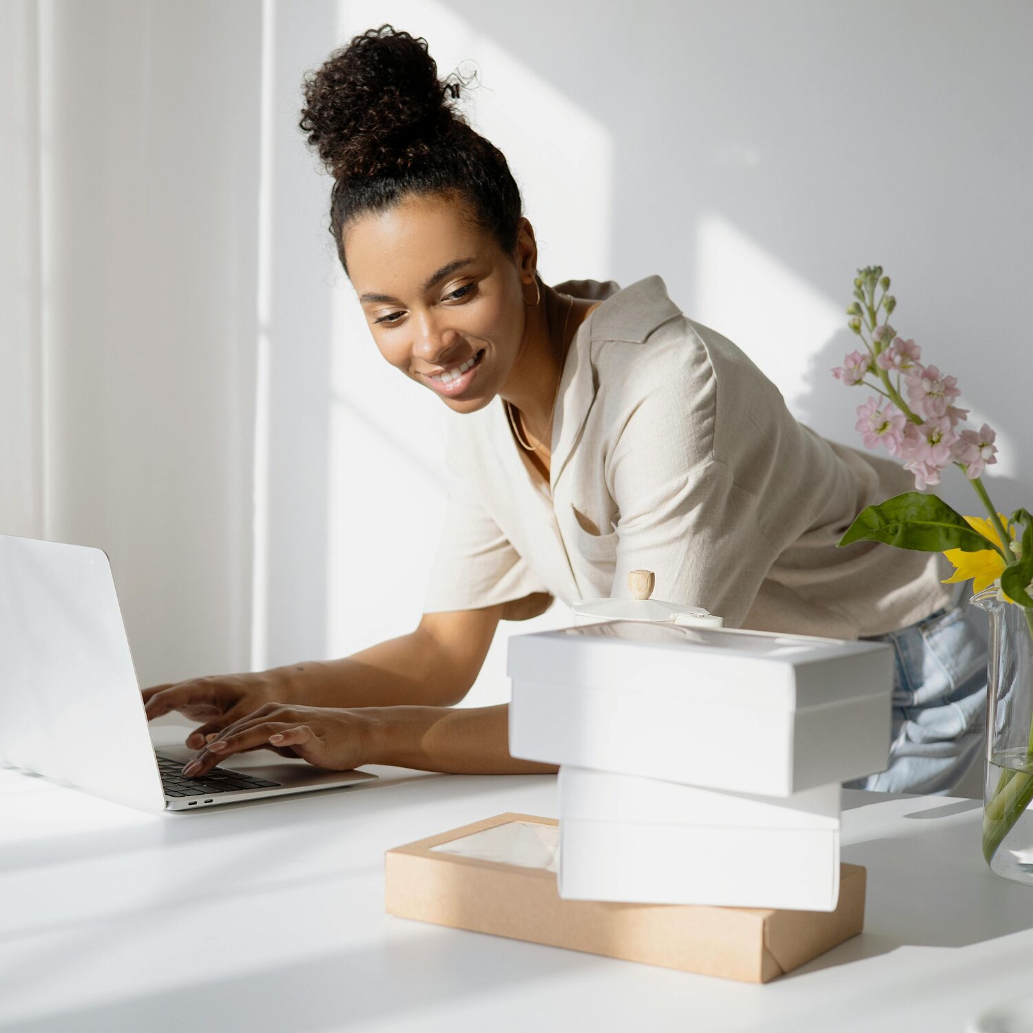 Young Black woman typing on her laptop, at a white desk with boxes on it.