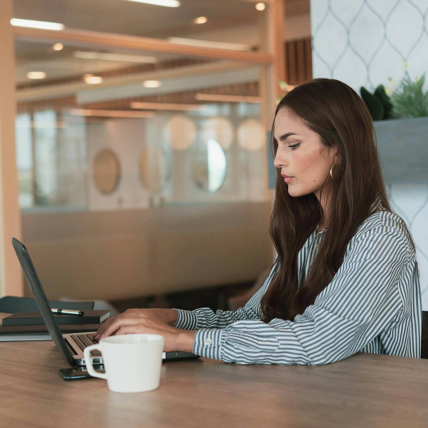 Young Latin woman in a blue and white pinstripe blouse, typing on a laptop, sitting at a wood desk.