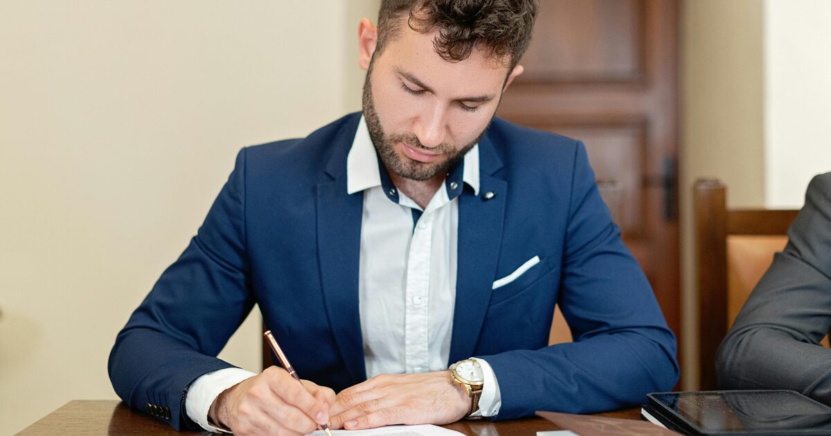 204 A Caucasian man sitting at a table, signing paperwork. He is wearing a navy tuxedo.