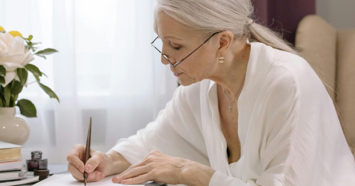 205 Senior Caucasian woman in a white dress, sitting at a dark brown table, signing a piece of paper.