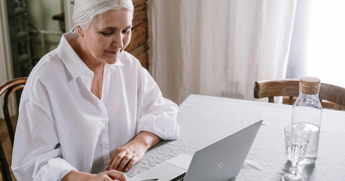 206 Senior Caucasian woman in a white shirt, sitting at a white table, reading something on her laptop.