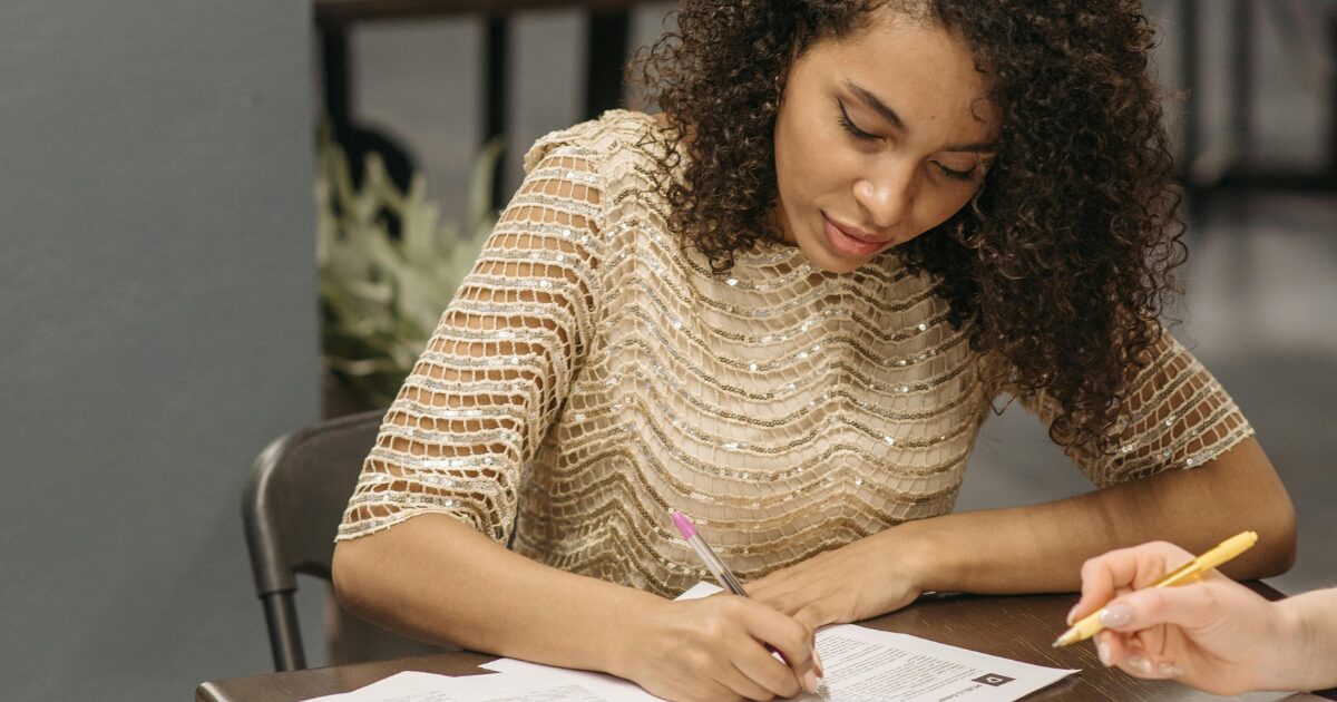 207 Black woman singing paperwork at a table, she is wearing a gold sparkly top.