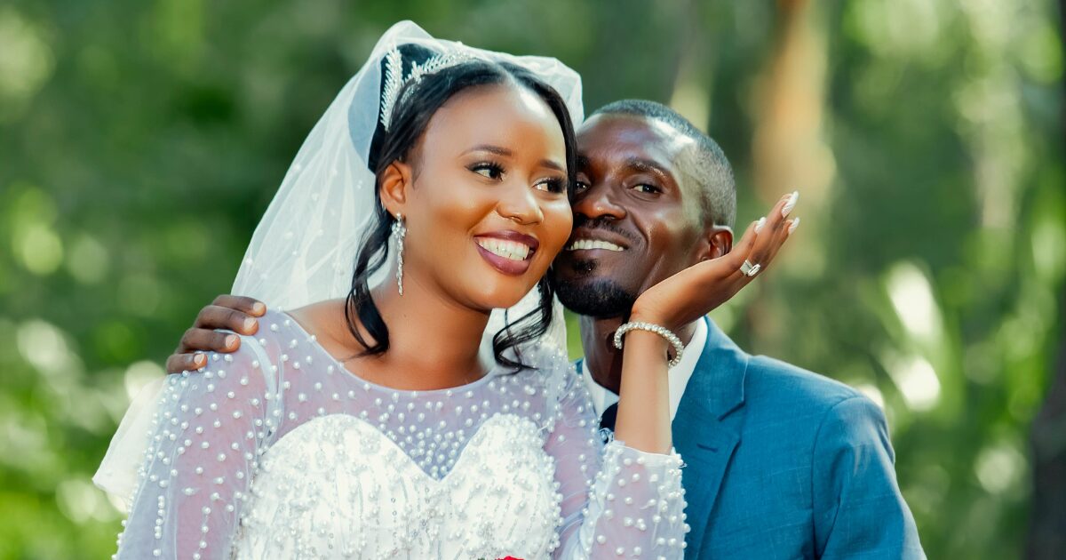 A Black woman wearing a white wedding dress with a man wearing a blue tuxedo. Both are smiling and he has his arm around her.