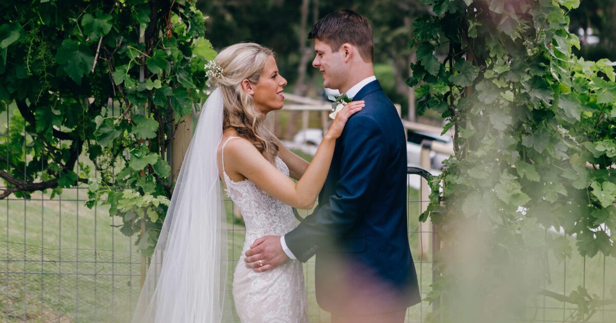 A blonde Caucasian woman wearing a wedding dress, standing at the alter with a Caucasian man wearing a navy tuxedo. She has her hands on his shoulder and he is holding her hips.
