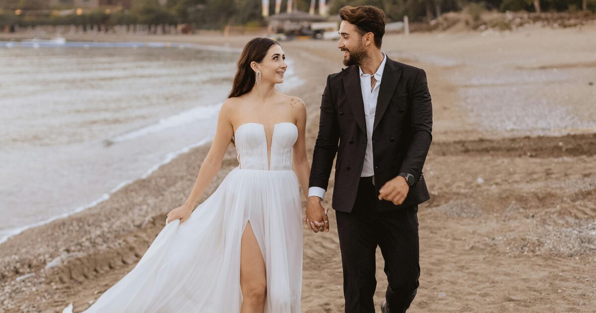 A Middle-Eastern woman in a wedding dress holding hands with a South Indian man in a tuxedo. They are walking on the beach.