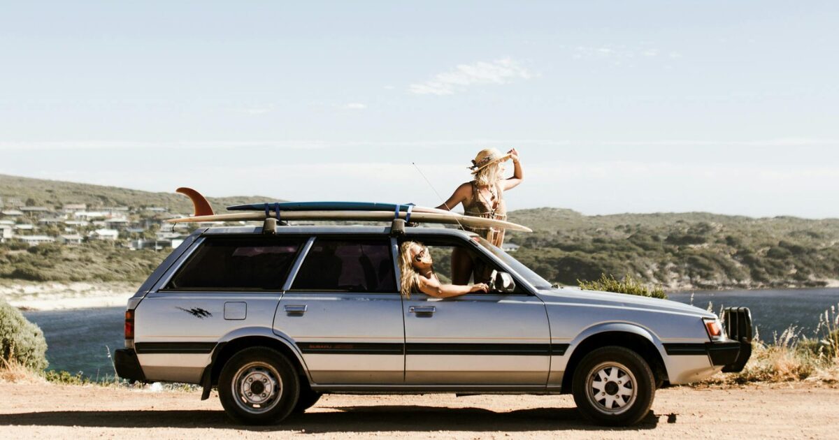 Two Caucasian young women in a silver vehicle, parked in front of a lake and mountains on a sunny day.