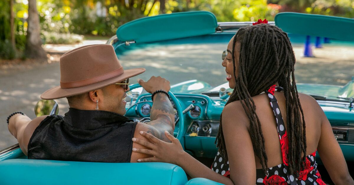A Black man driving a turquoise convertible, laughing with a Black woman in the passenger seat.