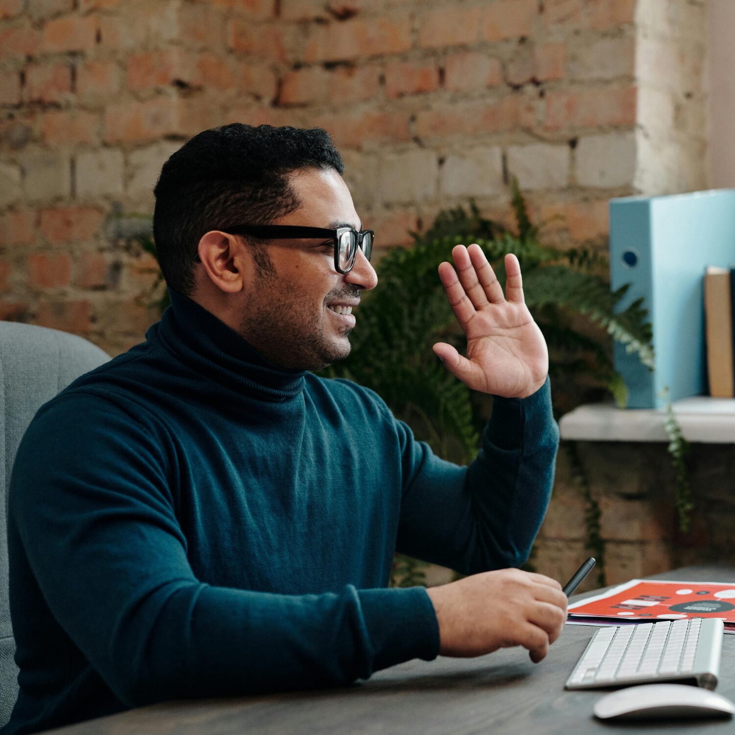 Adult Middle-Eastern man wearing a teal turtleneck and eyeglasses. He is holding his left hand up, doing the motion of giving an oath or affirmation.