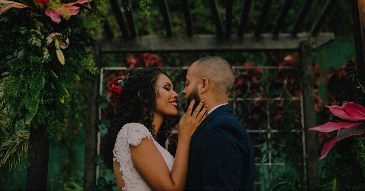A Black woman wearing a wedding dress standing in front of a Black man in a tuxedo. They are surrounded by tropical plants at an alter.