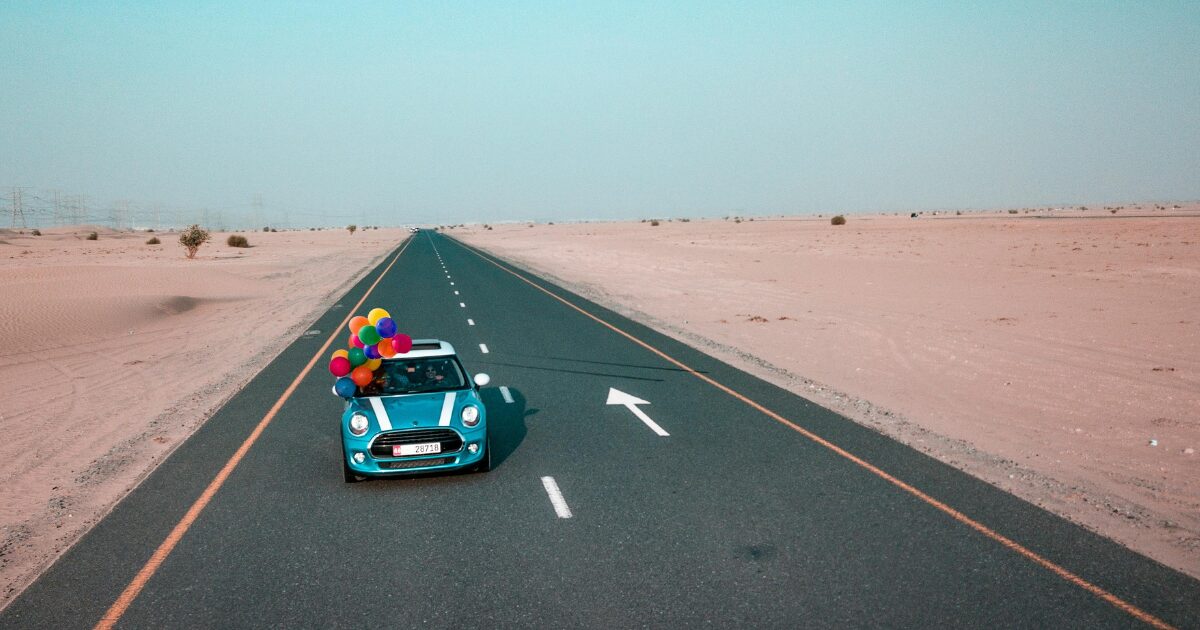 A blue car with colourful balloons attached to the top, driving down an open desert road.